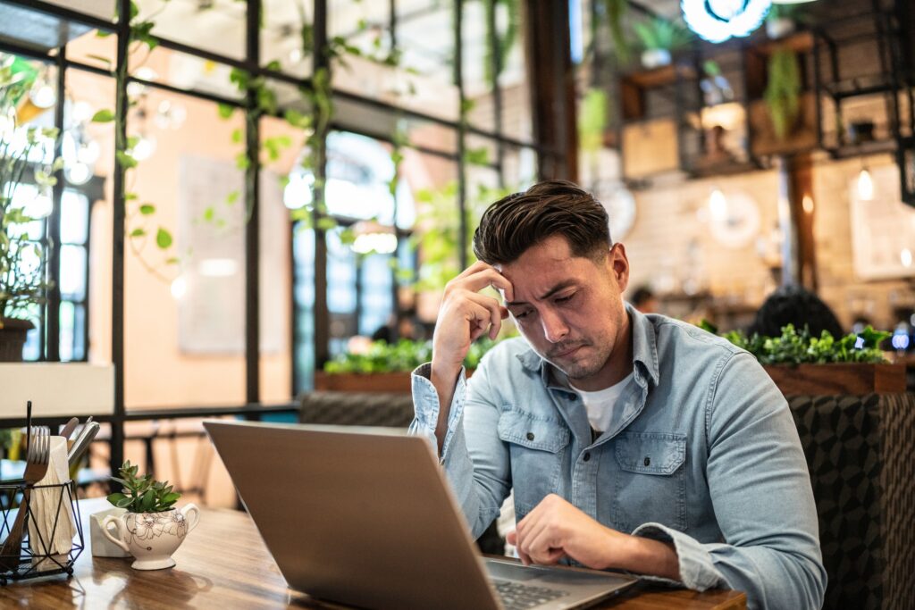 A man experiencing productivity anxiety while working on his laptop at a cafe.